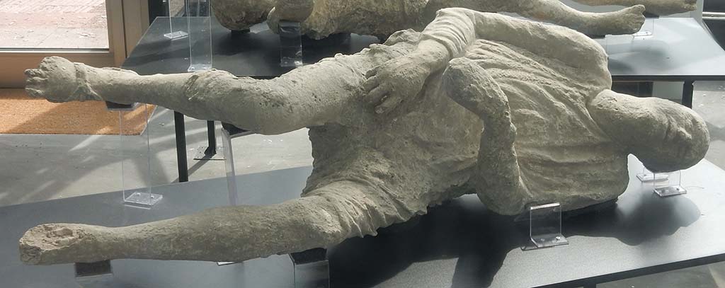Pompeii. May 2018. General view of two more plaster-casts. Photo courtesy of Buzz Ferebee.
In the foreground is the plaster-cast known as Victim 15, the heavily clothed man.
According to Notizie degli Scavi, (1890, P.128) the body of a man wrapped in his cloak and lying on his left side was found on 12th March 1890 in the excavations outside of the Stabian Gate. It was of a young man, of slender figure, lying on his left side, wound up in his cloak and wearing short pants which exposed his legs from above the knee. On his right foot could be clearly seen the sandal he was wearing.
See Dwyer, E., 2010. Pompeii’s Living Statues. Ann Arbor: Univ of Michigan Press. (p.107-8).
