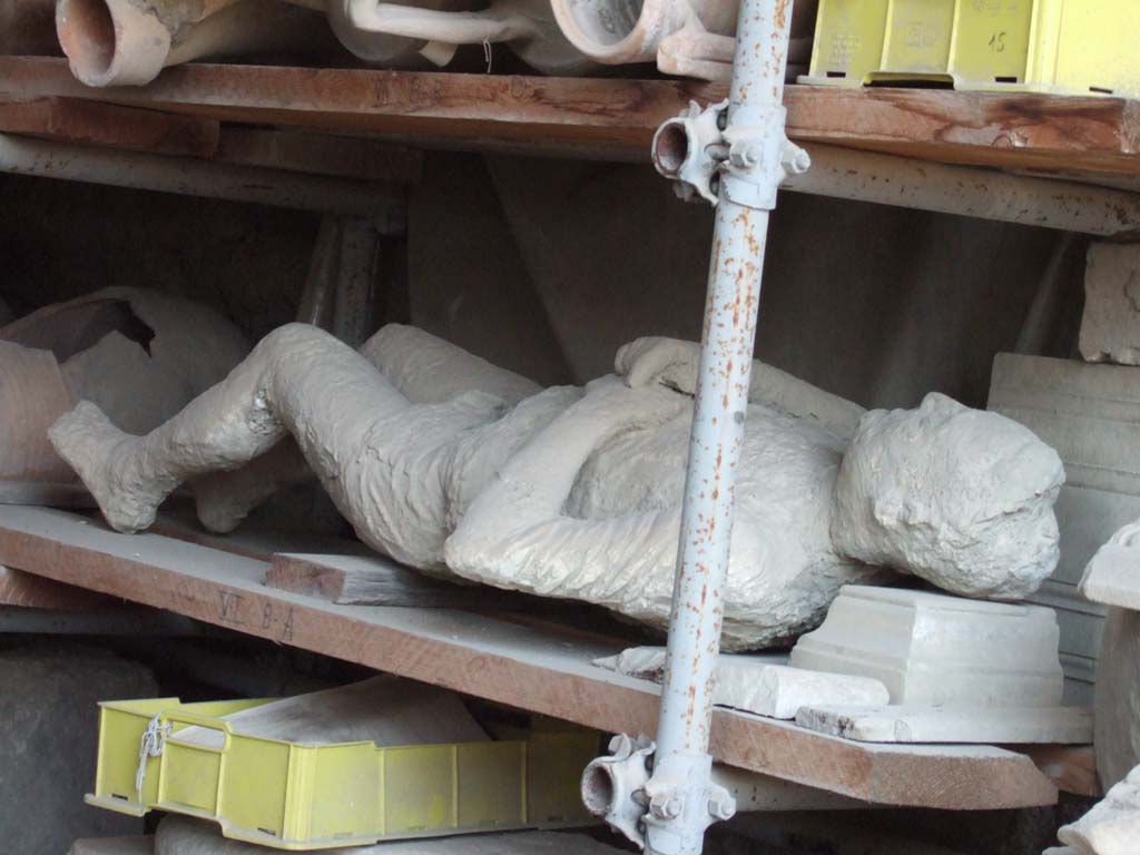 VII.7.29 Pompeii. May 2006. Forum Granary store. Plaster cast of body, known as victim number 13.
Garcia y Garcia described a mature man, already old, but perhaps still vigorous, fallen on his back, wrapped in a wide cape, with hands on his chest and with slightly drawn up legs.
He had a fine aquiline nose and a smile was on his lips. On the right leg was a circular ring that denoted a slave.
This plaster cast was obtained outside the Stabian Gate on the 11th of October 1889, at approximately 10 metres if you are exiting and turn to the right.
A plaster cast was also made of a woman who was found next to this man.
See Garcia y Garcia, L., 2006. Danni di guerra a Pompei. Rome: L’Erma di Bretschneider. (p. 193-4, Figg. 451-3).
According to Dwyer, this plaster cast was of victim number 14.
See Dwyer, E., 2010. Pompeii’s Living Statues. Univ. of Michigan Press: (p.103)
