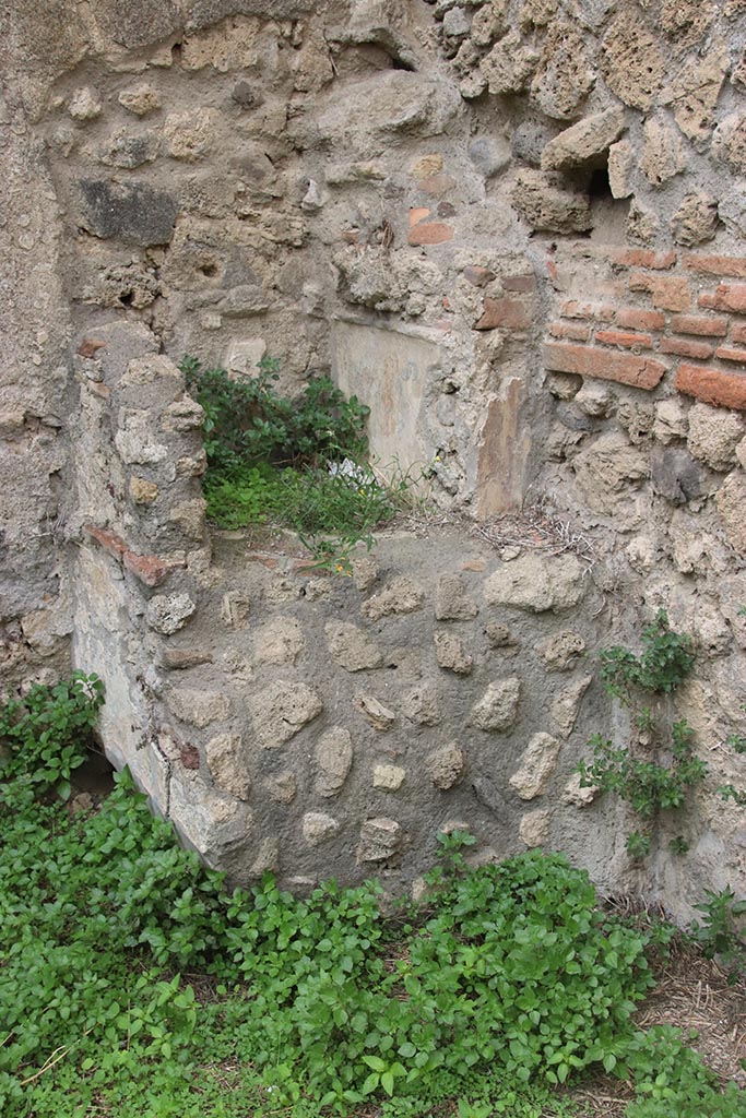 IX.7.12, 14 or 16 (?) Pompeii. October 2024.
Garden area “e”, lararium in north-west corner. Photo courtesy of Klaus Heese.
According to Boyce –
Against the west wall of the garden stands an aedicula, built upon a square masonry base (0.92 square, h.0.70).
The sides of the base are decorated with green plants painted on the white ground.
Above the base, walls enclose a niche (h.0.42, w.0.52, d.0.62).
In front of the structure stands a small altar (0.24 by 0.23, h.0.80).
The roof (no longer preserved) originally extended out over the altar as well as the niche, and the front of it was adorned with a pediment.
It was supported by two columns, the marks of the bases of which are still to be seen near the corners of the base.
On the walls within the niche is the lararium painting.
On the back wall to the right of an altar, stands the Genius pouring a libation upon it.
To the left of the same altar is a serpent, the head of which was done in stucco relief.
On each of the side walls are painted three figures: from back to front – a plant, a Lar, a man in a white garment holding in his left hand an object resembling a black cup, with a patera in his right hand.
He quotes references – Not. Scavi, 1881, 92-93; Bull. Inst., 1883, 146.
In Not. Scavi, it is described as House, only partly excavated – “and corresponds to, after the sixth doorway, west side, counting from the north-west”.
In Bull. Inst, it is described as “West side, first entrance counting from the south, garden in part excavated.”
See Boyce G. K., 1937. Corpus of the Lararia of Pompeii. Rome: MAAR 14. (p.89, no.445).