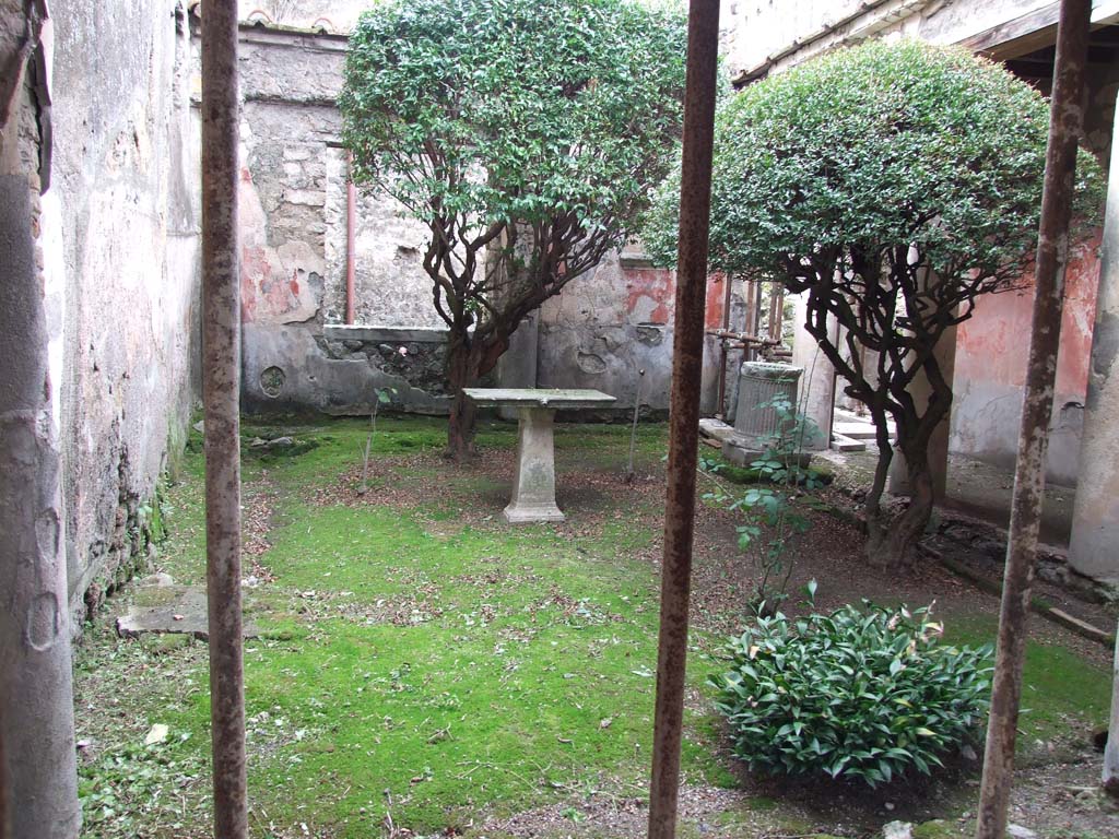 I.7.7 Pompeii. December 2006. Pseudoperistyle, looking west from east portico.
According to Jashemski, the garden was enclosed on the north and east by a portico supported by five columns and two engaged columns. 
The room on the west side had a large window looking into the garden. 
Maiuri pointed out that the large tree-root found against the south wall showed that the little garden had been well shaded.
The nail holes in the wall indicated that a vine had been carefully trained up the wall.  
The tree-root cast can be seen half way up the garden next to the wall on the left (0.80 x o.80m at ground level).  
There was a puteal over the cistern opening in the north-west part of the peristyle.  
Another puteal was found in the garden.  
The table (cartibulum) in the garden was decorated with lion heads on one side. 
The small circular marble base probably held a statuette. 
The arched niche with an aedicula façade on the west wall was a Lararium.
See Jashemski, W. F., 1993. The Gardens of Pompeii, Volume II: Appendices. New York: Caratzas. (p.37)
