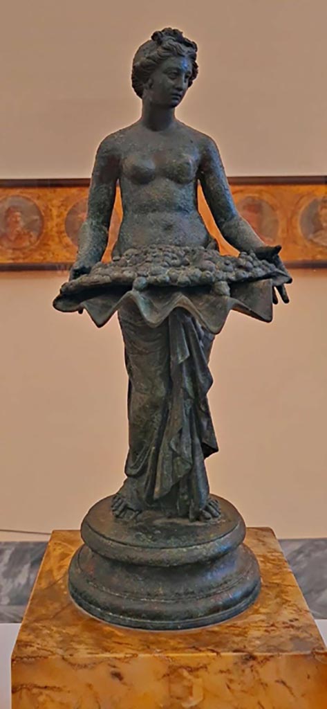 I.7.12 Pompeii. October 2023.
Bronze statuette of Pomona. Photo courtesy of Giuseppe Ciaramella.
On display in “L’altra MANN” exhibition, October 2023, at Naples Archaeological Museum.
According to Jashemski, this statue can be seen in her photo on p.39, fig.38, inv. 144276.
See Jashemski, W. F., 1993. The Gardens of Pompeii, Volume II: Appendices. New York: Caratzas, (p.38).
According to Naples Museum –
“This bronze statue, with another of a bathing Aphrodite, was described as Bathing Aphrodite with Nymphs, inv. S.N.”
The description card described the group as from the Vesuvian area, as opposed to I.7.12, Pompeii.
So the bronze statue may, or may not, come from I.7.12.