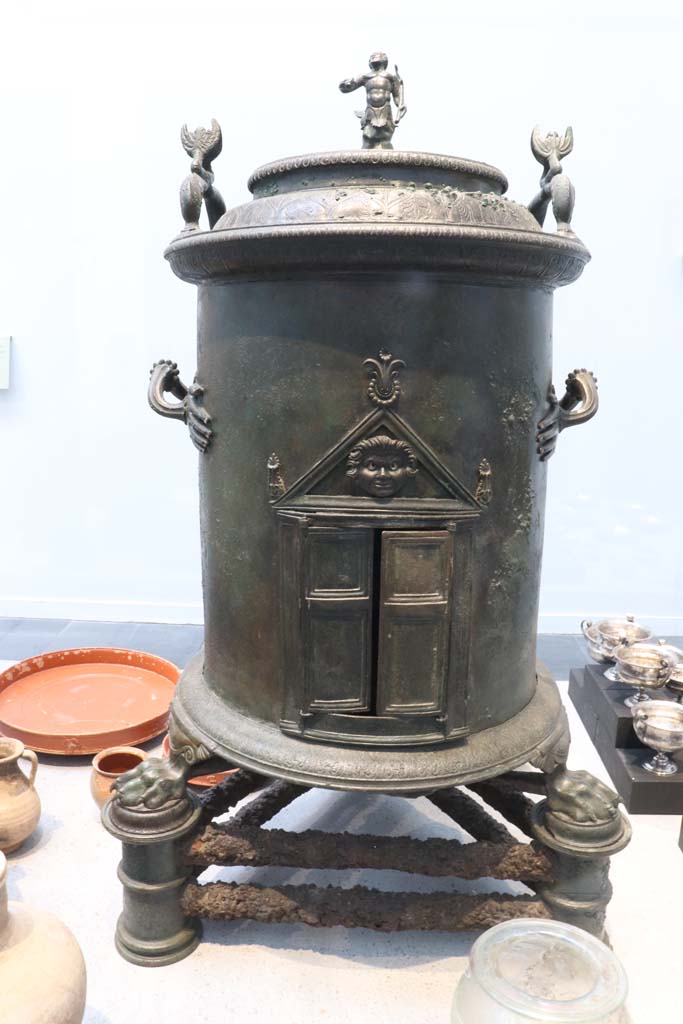 VIII.1.4 Pompeii. February 2021.
Bronze cylindrical food warmer found under the stairs in the servant’s quarters of I.8.17.
On display in Antiquarium. Photo courtesy of Fabien Bièvre-Perrin (CC BY-NC-SA).
According to the Musée Maillol, the Romans created a range of richly decorated devices, usually in bronze, for storing hot water during triclinium banquets.
The hollow cylinder of the latter rests on an iron tripod.
The feet are in the form of lion's paws, the handles end in small hands.
A small temple with a Gorgon in the tympanum can be seen in the opening.
Two dolphins and a Triton decorate the lid.
See Le Musée Maillol : Pompéi - Un art de vivre, l’exposition du 21 septembre 2011 au 12 février 2012. Communiqué de presse.
Parco Archeologico Pompei, inventory number 6798.