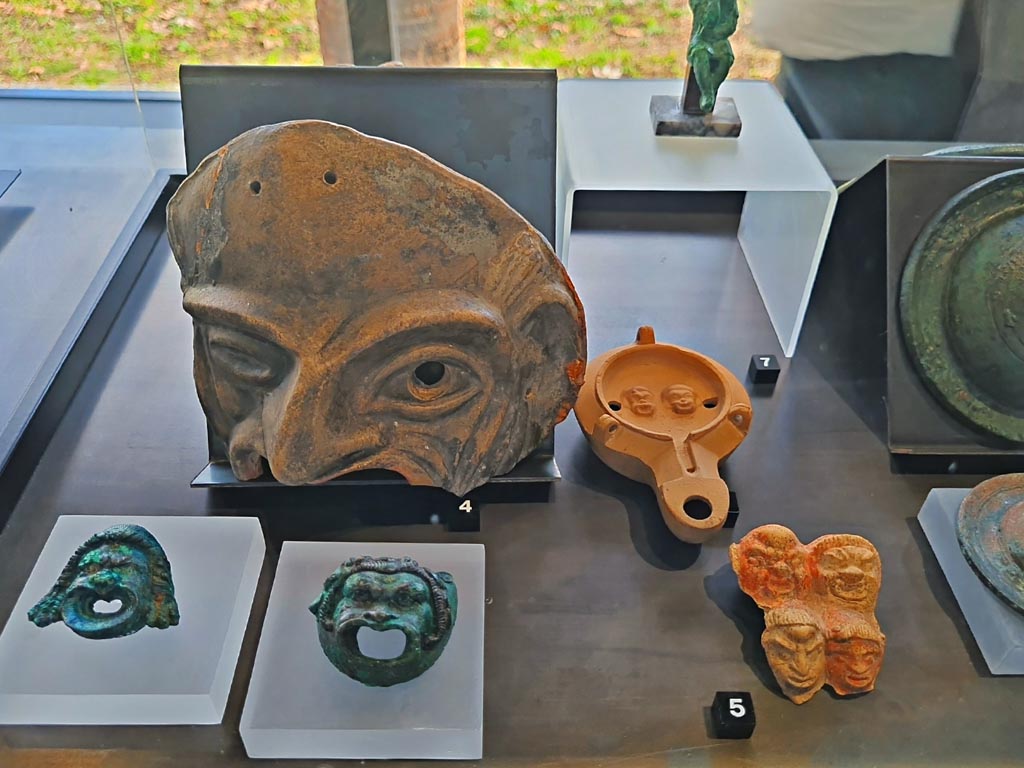 I.9.13/14 Pompeii. March 2024. No.5 – Plaque with theatre masks, (Ist century BC – Ist century AD). Photo courtesy of Giuseppe Ciaramella.
On display in exhibition in Palaestra entitled – “L’altra Pompei, vite comuni all’ombra del Vesuvio”.
No.3, lower left – Bronze wall sconces with theatre masks, provenance not known, but from Pompeii. (Ist century AD).
No.4 – Terracotta theatre masks Pompeii – provenance not known, but from Pompeii. (Ist century AD).
No.6 (number hidden, but beneath No.7) – Oil lamp with theatre masks, from I.9.9.