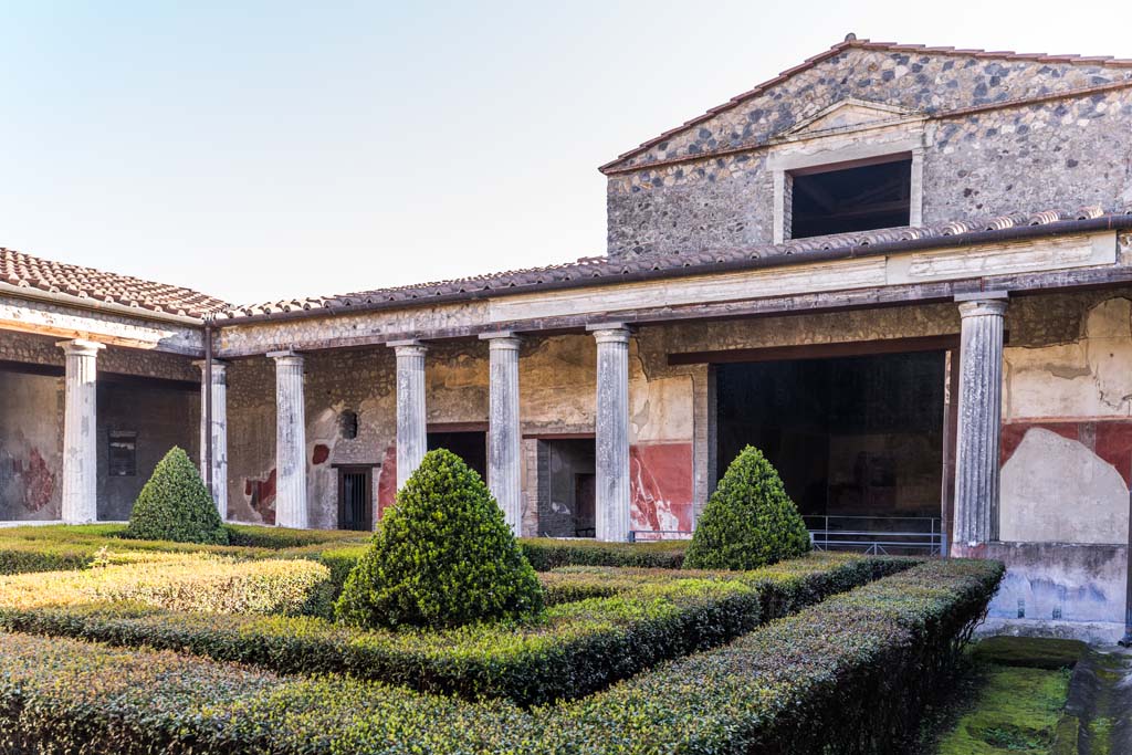 I.10.4 Pompeii. April 2022.
Looking across peristyle garden towards east side from south-west portico.
The pediment and window above room 18 can be seen. Photo courtesy of Johannes Eber.