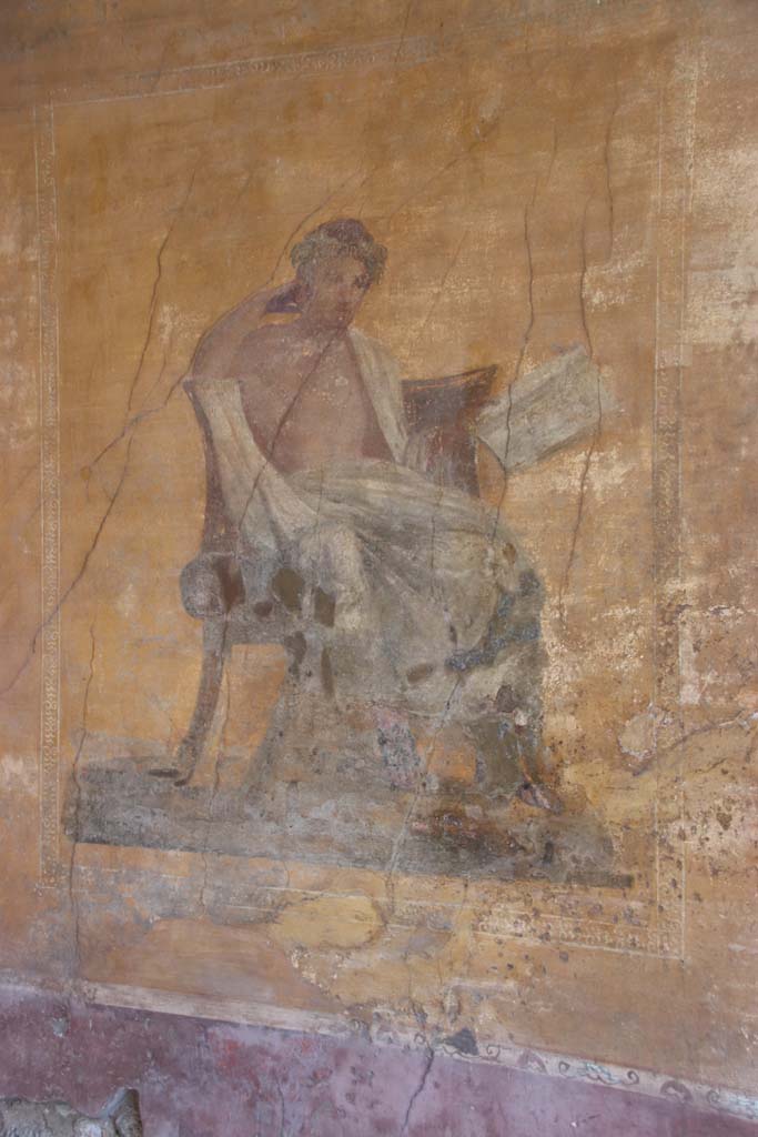 I.10.4 Pompeii. September 2021.
Alcove 23, west wall with wall painting of a seated poet. Photo courtesy of Klaus Heese.
The name of Menander was inscribed by the painter on the skirt of the man’s cloak.
In this alcove there are also painted masks, with possibly Bacchus sitting on a chair with a robe covering his legs, and a painted table.