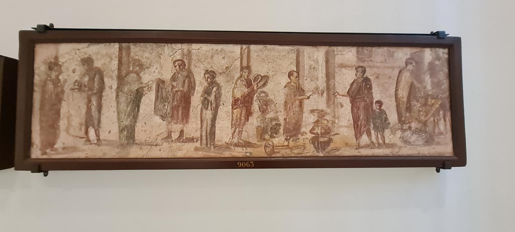 II.4.3 Pompeii. April 2023. Paintings of the Forum frieze, on display in “Campania Romana” gallery in Naples Archaeological Museum.
Above (inv. 9063) – Vendita di tessuti, pentolame e pane davanti a colonnato.
(Sale of fabrics, pots and pans and bread in front of a colonnade.) Photo courtesy of Giuseppe Ciaramella.