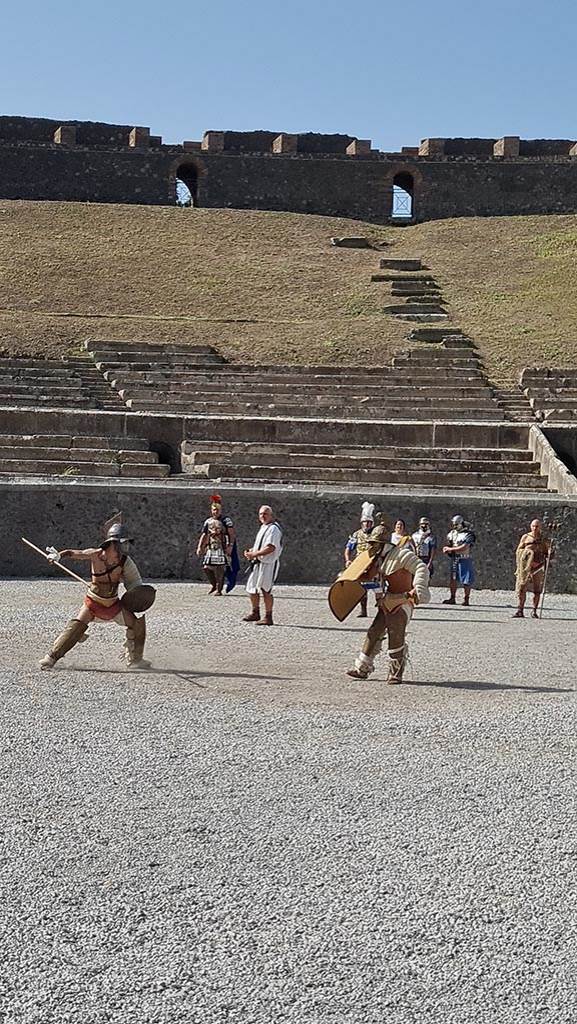 II.6 Pompeii. September 2024.
Looking west across amphitheatre towards Gladiators in combat rehearsing.
Photo courtesy of Giuseppe Ciaramella.