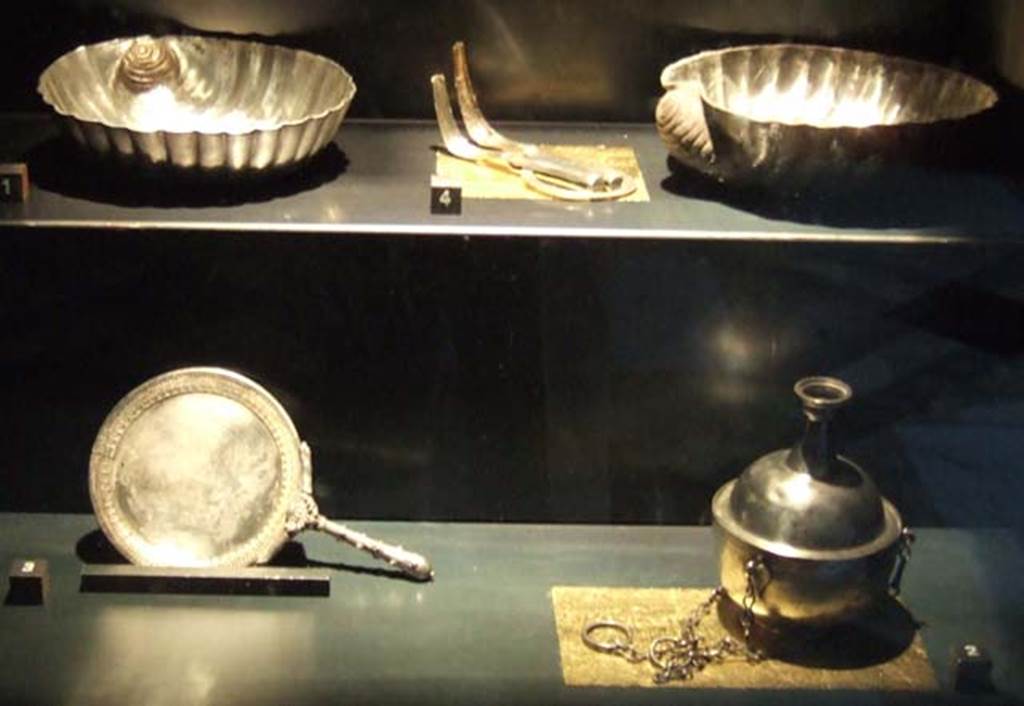 V.1.18 Pompeii. Silver found in the ala “e”.
Now in Naples Archaeological Museum.
Top left to bottom right:-
Two shell pattern containers. Inventory numbers 110863 and 110864
Between them are two strigili joined by a ring. Inventory number 110862.
Concave circular mirror with handle. Inventory number 110861.
Container with cylindrical neck. Inventory number 110841.