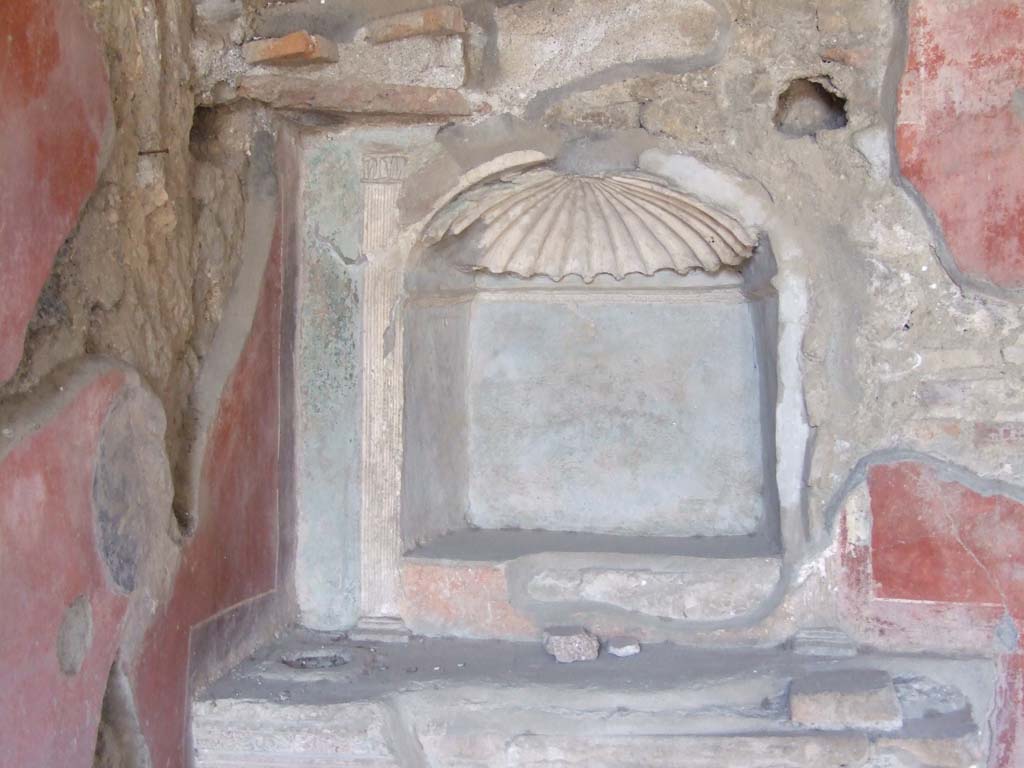 V.3.4 Pompeii. March 2009. Lararium. Blue painted arched niche with shell crowned top and modelled internal stucco cornice.
The lararium originally had a roof with pediment, the remains of which can be seen above the shell.
The pediment was supported by two columns, the marks of which can be seen on the top of the podium.
Around the outside curve of the niche was a stucco cornice with an egg pattern.
See Boyce G. K., 1937. Corpus of the Lararia of Pompeii. Rome: MAAR 14. (p. 38, 111, Pl 38,1).