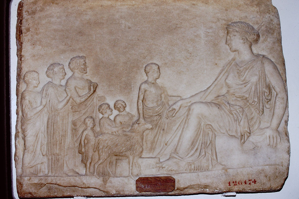 V.3.10 Pompeii. 1984. Marble relief showing a goddess, most likely Aphrodite, enthroned on a rock.
Found on 12th September 1901, in the small garden, not in situ but leaning against the western wall.
Marble tablet 475mm high, 600mm wide and 45mm thick.
See Notizie degli Scavi di Antichità, 1901, p. 400f.
On the left, in front of the goddess, is a group of two women, a man, three children and an altar servant leading a sacrificial ram.
Now in the Naples Archaeological Museum, inventory number 126174.
According to Kraus, this relief was found leaning against a wall in the small garden, perhaps waiting to be set into the wall.
See Kraus T. and von Matt L., 1975. Pompeii and Herculaneum: Living cities of the dead. New York: Abrams. (p.193, fig 266
Source: The Wilhelmina and Stanley A. Jashemski archive in the University of Maryland Library, Special Collections (See collection page) and made available under the Creative Commons Attribution-Non Commercial License v.4. See Licence and use details.
J84f0012