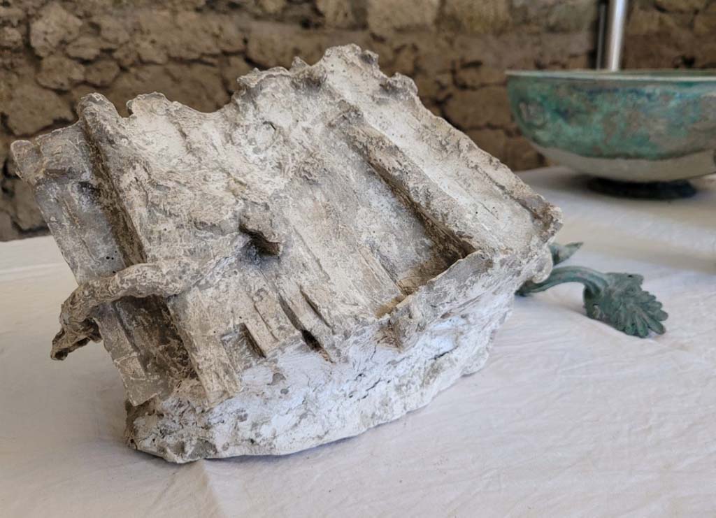 V.3.13 Pompeii. 2022. Upper floor.
The upper rooms were excavated first, however the materials discovered were primarily found where they had fallen into the area of the rooms below.
Amongst these, of great documentary value is the small cast of the waxed tablets. A truly unique find, it has allowed us to create the first example of a cast which allows a perfect yield of both volume and detail. It is a group of seven triptychs, tied together by a small cord both horizontally and vertically. The polyptych had probably been stored on a shelf, together with other ceramic and bronze objects.
Discovered within a large cupboard which collapsed during the eruption were various other well-preserved ceramic vessels for everyday use in the kitchen and the canteen, but also vessels in sigillata (a type of Roman fine table ceramic) and in glass. Alongside this was a set of bronze vessels, among which a well-preserved pelvis (bowl) with a beaded base and handles with palmette attachments particularly stands out. There are in addition two bronze jugs, one of which features a handle with a sphinx-shaped attachment above and a lion’s head attachment below.
Gli ambienti superiori sono stati scavati per prima, tuttavia i materiali rinvenuti sono stati prevalentemente ritrovati in fase di caduta nella volumetria degli ambienti sottostanti.
Tra questi di gran valore documentario è il piccolo calco delle tavolette cerate. Un unicum per la tipologia di ritrovamento, che ne ha permesso di realizzare il primo esemplare di calco, che ne consente la perfetta restituzione della volumetria e dei dettagli. Si tratta di un gruppo di sette trittici, legati tra essi da un cordino sia in senso orizzontale che verticale. Il polittico doveva probabilmente essere conservato su qualche scaffale, unitamente ad altri oggetti in ceramica e in bronzo.
Contenute all’interno di un grosso armadio, crollato durante l’eruzione, sono inoltre state recuperate diverse forme ceramiche d’uso comune, da cucina e da mensa, ma anche forme in sigillata (tipo di ceramica romana fine da mensa) ed in vetro, molto ben conservate. A queste si affianca un piccolo set di forme in bronzo, tra cui spicca una ben conservata pelvis (bacile) con fondo perlinato ed anse con attacchi a palmette. Con essa anche due brocche bronzee, una delle quali con ansa con applique sormontante a forma di sfinge ed attacco inferiore a testa leonina.
Photograph © Parco Archeologico di Pompei.