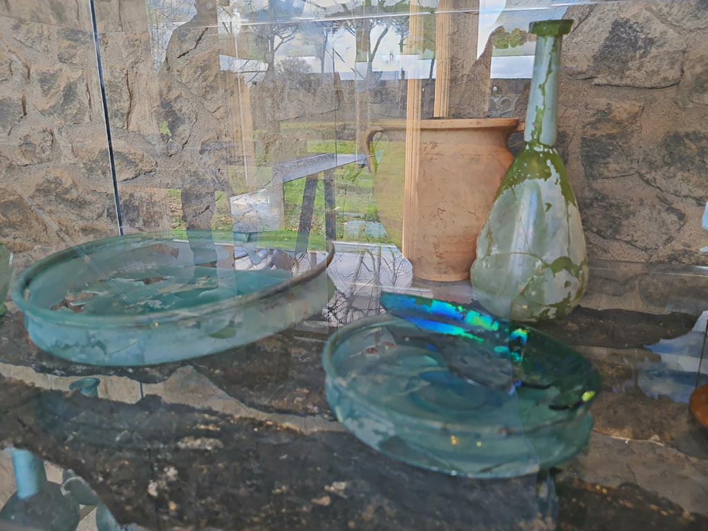 V.3.13 Pompeii. March 2024. Restored glass items and carbonised wooden shelf in reconstruction of a cupboard/cabinet.
Oggetti in vetro restaurati e mensola in legno carbonizzato nella ricostruzione di un armadio/armadio.
Photo courtesy of Giuseppe Ciaramella.
On display in exhibition in Palaestra entitled “L’altra Pompei, vite comuni all’ombra del Vesuvio”.