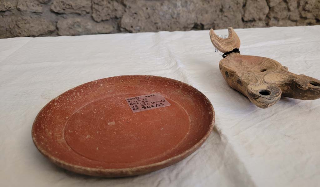 V.3.13 Pompeii. 2022. Room 3b, bedroom. Chest contents.
A small sigillata plate and a double-spouted lantern with a bas-relief depiction of the transformation of Zeus into an eagle.
Un piattino in sigillata (tipo di ceramica romana fine da mensa) e una lucerna a doppio beccuccio con bassorilievo raffigurante la trasformazione di Zeus in aquila.
Photograph © Parco Archeologico di Pompei.