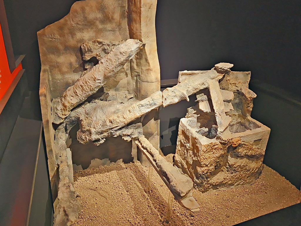 V.3.13 Pompeii. March 2024.
Room 3b, on the left is the cast of the remains of a bed with a headboard to which a pillow is attached.
On the right is a copy of an open wooden trunk, on which the beams and boards of the floor above have collapsed.
Next to it, is a round three-legged table (see photo above this one).
On display in exhibition in Palaestra entitled “L’altra Pompei, vite comuni all’ombra del Vesuvio”. Photo courtesy of Giuseppe Ciaramella.