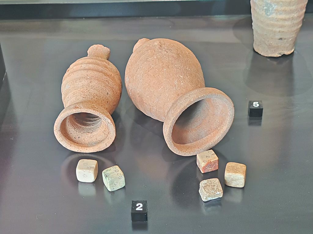 V.4.13, Pompeii. March 2024. No.2 – Worked bone dice. Photo courtesy of Giuseppe Ciaramella.
On display in exhibition in Palaestra entitled – “L’altra Pompei, vite comuni all’ombra del Vesuvio”.
Perhaps these are the ones mentioned by Sogliano –
“Room Q – leaning against the western wall was a chest or cupboard, which was found on 6th April 1903………………..
The following items were stored there: ………………… Bone: three dice and five checkers.”
See Notizie degli Scavi di Antichità, 1905, (page 92-93).