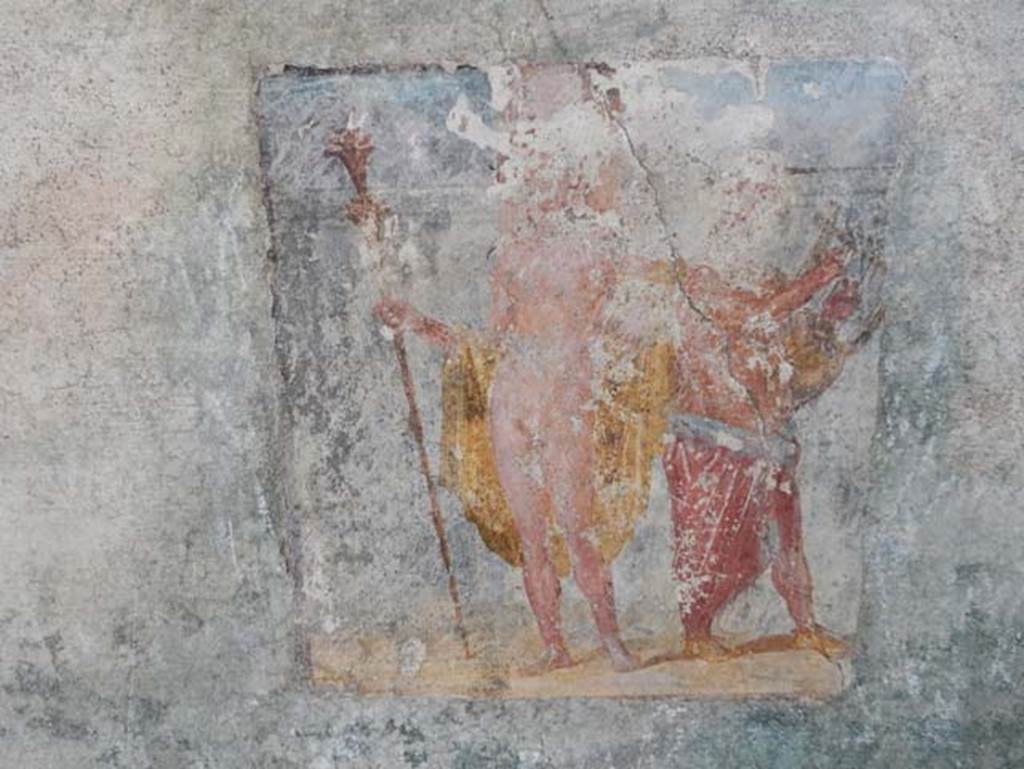 V.4.a Pompeii. May 2015. Room ‘s’, wall painting of Dionysus (Bacchus) accompanied by Silenus playing the lyre.
From the east wall of the summer triclinium. Photo courtesy of Buzz Ferebee.
According to Kuivalainen, who describes the painting as –
“A composition of two standing figures on yellowish terrain against a simple architectural background.
An effeminate youth with fair complexion stands frontally, with his weight on his right foot; in his right hand he holds a thyrsus with a bunch of leaves and a torch-like decoration on the top; ……..”
Kuivalainen then comments –
“Though the pose is customary to practically naked Bacchus leaning on a playing Silenus, the figure would possibly be a hermaphrodite, not Bacchus. This painting had a parallel in room ‘t’ of the House of Caecilius Iuncundus (V.1.26, our room 22). The torch-like thyrsus would be a rarity to Bacchus. It is more common for hermaphrodites and divinities of light.”
See Kuivalainen, I., 2021. The Portrayal of Pompeian Bacchus. Commentationes Humanarum Litterarum 140. Helsinki: Finnish Society of Sciences and Letters, (F14, p.170).