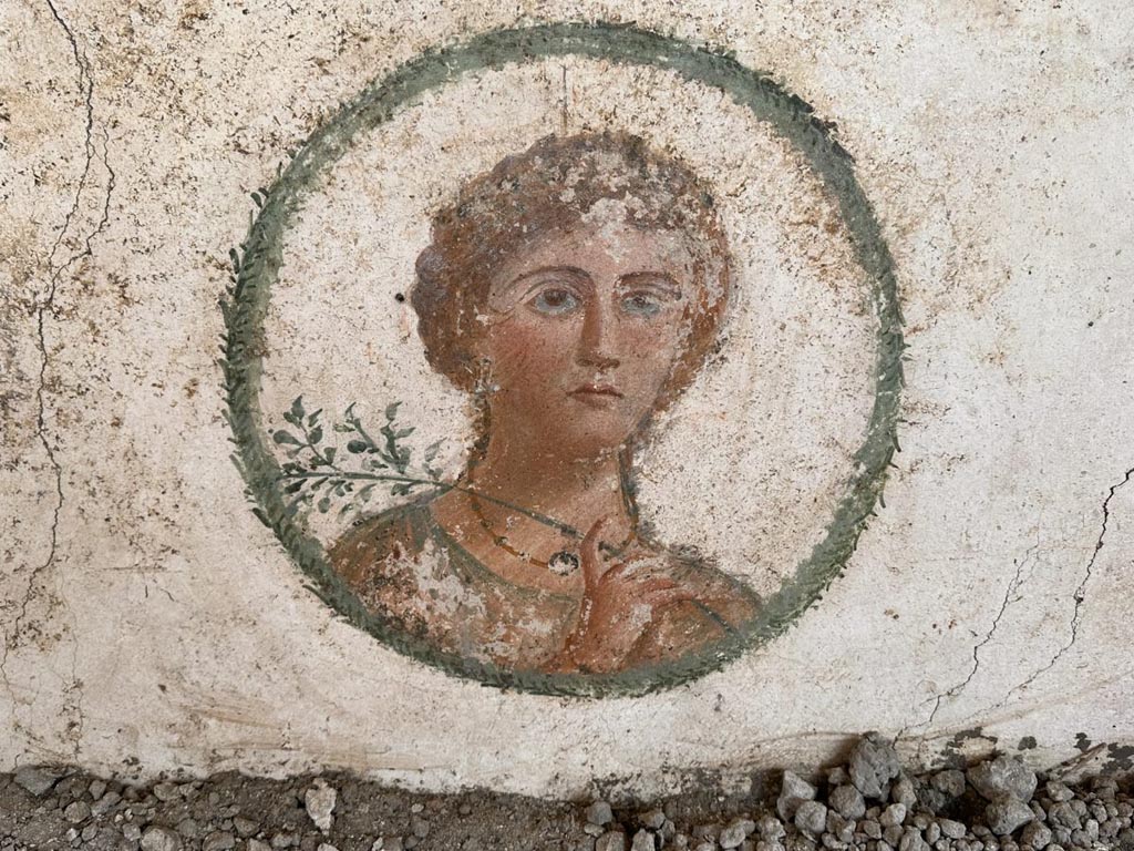 V.6.12 Pompeii. March 2024. Amb. 7. Portrait medallion of a female figure adorned with jewels and holding an olive branch.
On the central panel of each wall there is a clipeus with a female portrait, two of which have already emerged in 2018: one captured in a pensive attitude; the other, with a veiled head, is characterized by a twig. The ongoing investigation has revealed two further portraits of great refinement: a female figure adorned with jewels and characterized by the presence of an olive branch; the other characterized by a crown of vine leaves around the head.
Vedi/See E-Journal 09: 21.12.23, Scavo e restauro della Casa di Leda – Regio V, 6. Nuovi rinvenimenti, p. 3, Fig. 3.
Photograph © Parco Archeologico di Pompei.