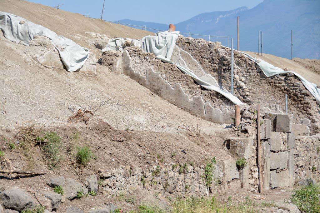 V.6.15 and V.6.14, Pompeii. 2018. Looking south along front facades on east side of Via del Vesuvio during works.
Photo © Parco Archeologico di Pompei.
(c.2018-2019). In order to improve the drainage and security for visitors to the site, the soil, ash and exuberant vegetation was removed from the slope of the unexcavated land on the east side of Via del Vesuvio (Insula V.6).
This has cleared the area to enable the street boundary wall and rooms at its immediate rear to be viewed from Via del Vesuvio.