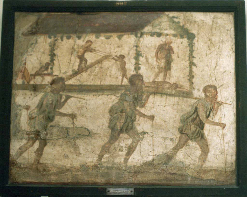 VI.7.9 Pompeii. Wall painting of the Procession of the Carpenters.
Originally found on pilaster between entrances VI.7.8 and VI.7.9.
Now in Naples Archaeological Museum. Inventory number 8991.
See Helbig, W., 1868. Wandgemälde der vom Vesuv verschütteten Städte Campaniens. Leipzig: Breitkopf und Härtel. (1480).
According to Leach, figures of Minerva, Mercury and Daedalus painted on its exterior pilasters (6.7.8-12), as well as another painting of a procession advertising the craft of Daedalus under the protection of Minerva and Mercury, were taken by Mau as an indication that this was a Carpenter’s workshop.
The sign showed 3 carpenters bearing a ferculum that included a figure of Daedalus and some workmen performing carpenters’ tasks.
A dead figure lies before Daedalus’s feet: this may well be a reference to the nephew Perdix whom the legendary artisan murdered through jealousy for his invention of the rake.
(PPM 4, p389-91, however, suggested that the sign indicated a perfumer’s shop that dealt in spices requisite to funeral rituals).
See Leach, E.W: The Social Life of Painting in Ancient Rome and on the Bay of Naples.