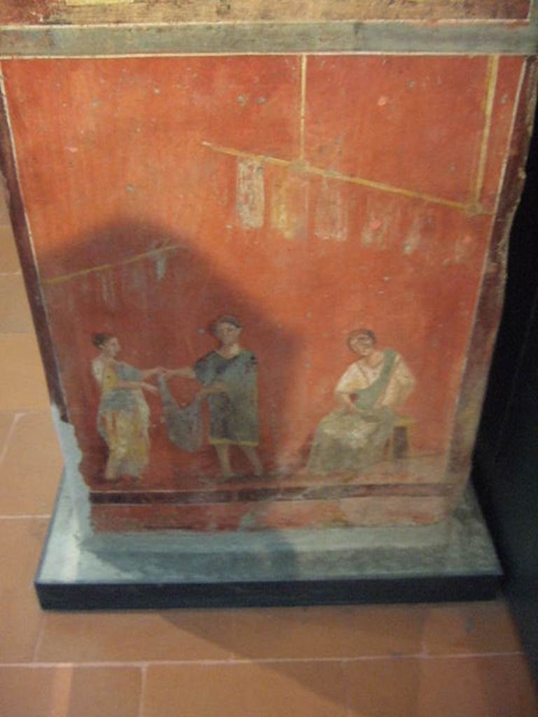 Pillar found in the Fullonica at VI.8.20. Painting of a woman collecting a garment, several of which are seen suspended on high on sticks. A woman, sitting, is cleaning the instrument for carding. Now in Naples Archaeological Museum. Inventory number 9974.