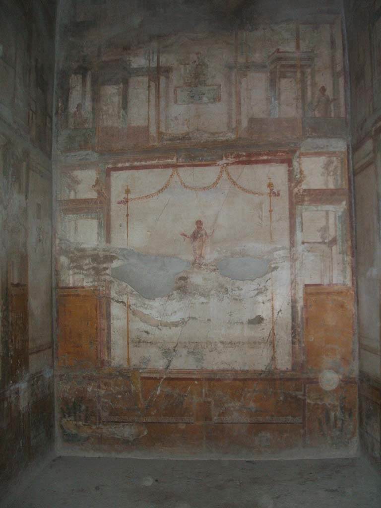 VI.8.23 Pompeii. May 2005. North wall of ala, with a yellow zoccolo (dado) and middle and upper areas painted white.
According to PPM, green painted plants could be seen in the narrow side panels of the zoccolo: in the central area was a rectangular area with garlands and a horizontal border of a “carpet” design on which peacocks rested.
Painted in the central aedicula was a female flying figure wearing a garment and cloak, outstretching her arms and holding a palm branch in the left hand.
In the upper part were architectural views with garlands, candelabra, shields and temples.
Painted in a square in the centre, was a clothed goddess on a throne, with sceptre in left arm and plate in the right hand.