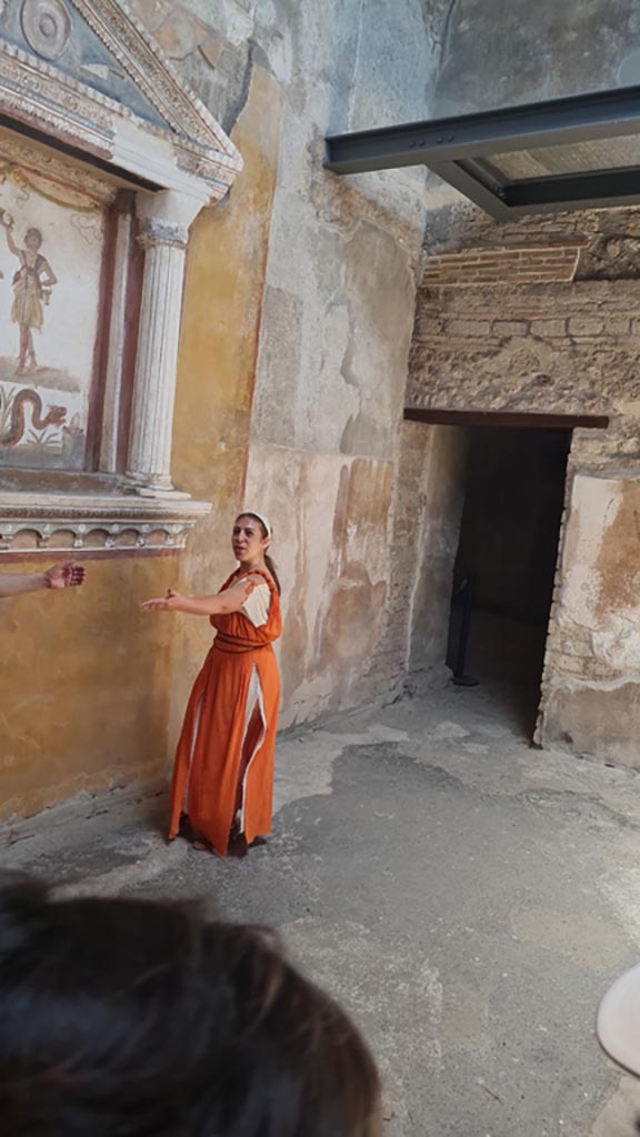 VI.15.1 Pompeii. 8th June 2024.
Looking towards north along west side of small atrium, with doorway to kitchen (w), on left.
Historical reconstruction entitled L’altra Pompei prende vita (The other Pompeii comes to life).