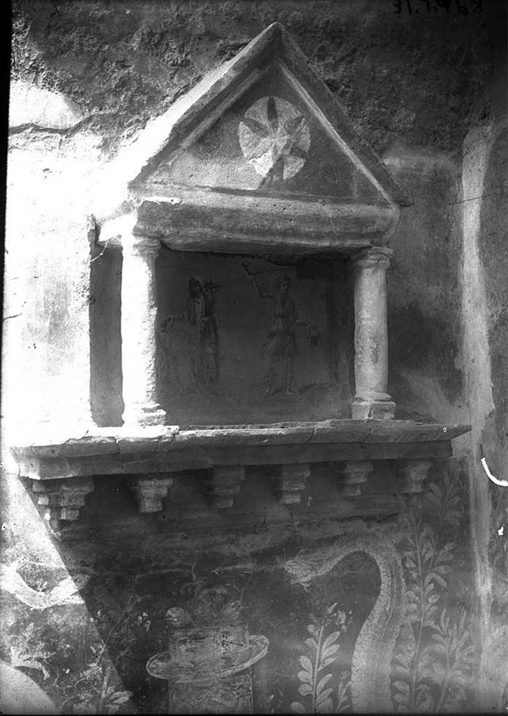 VI.15.23 Pompeii. Lararium on the west wall, in the north-west corner of the kitchen.
DAIR 31.2468. Photo © Deutsches Archäologisches Institut, Abteilung Rom, Arkiv.
According to Boyce, the whole was painted red except for a star on a white ground in the centre of the tympanum.
The curved back wall of the niche was painted blue, and upon it was the lararium painting.
On each side was a Lar, between them stood the Genius.
On the wall on each side of the aedicula was painted a garland and a flying bird.
Two large yellow serpents were painted on the wall beneath the aedicula and extended over onto the north wall on the one side.
On the other side, extending to the adjacent side of the hearth.
The serpent on the right had the larger body, a crest and a beard, the one on the left was smaller with no crest or beard.
They were on either side of a cylindrical altar painted in imitation of yellow marble beneath the aedicula.
The altar was furnished with two eggs and fruit.
See Boyce G. K., 1937. Corpus of the Lararia of Pompeii. Rome: MAAR 14. (p.56, no.219, and Pl.15, 1 and 2)
See Giacobello, F., 2008. Larari Pompeiani: Iconografia e culto dei Lari in ambito domestico. Milano: LED Edizioni. (p.183)
