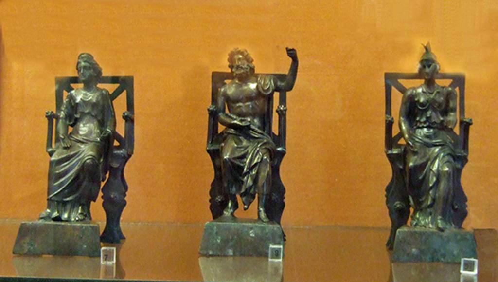 Three small bronze statues seated on thrones, found on the top step of the lararium of VI.16.7.
Juno (left), Jupiter in the centre and Minerva (right).
On the same step, to the left of Juno, stood the statue of Mercury.
The two Lares were placed, on either side of the group, but on the lower step.
Now in Naples Archaeological Museum. Jupiter is inventory number 133323, Juno is 133324 and Minerva is 133325.
See Boyce G. K., 1937. Corpus of the Lararia of Pompeii. Rome: MAAR 14, pp. 57-8, no. 221, and pl.38, 2
