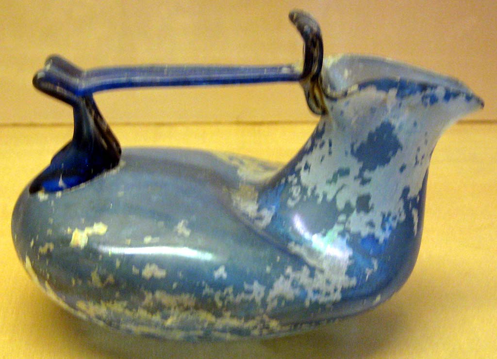 VI.16.10 Pompeii. Deep blue glass askos with encrustation found here.
It has a trilobate mouth, rigid handle with flat ribbon with rounded apex, lower bifurcated graft,
Now in Naples Archaeological Museum. Inventory number 133275.