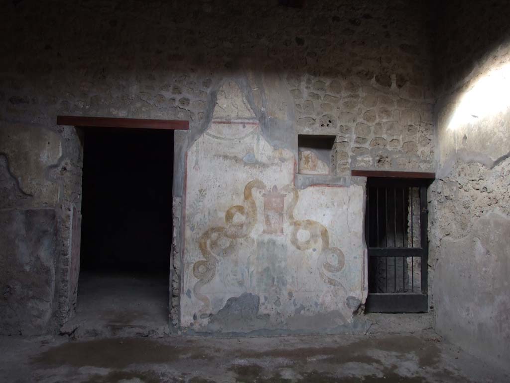 VI.16.15 Pompeii. December 2006. North wall of atrium with lararium between doorways to room I (left) and rooms K to M (right).
According to Boyce, the space between the two doorways on the north side of the atrium was painted with white stucco, bordered with red stripes.
This formed the large panel for the lararium painting.
A square niche was in the upper right-hand corner, on the rear wall of which was painted the figure of the Genius.
Hanging down on each side of him was a garland, on the side walls were painted red flowers with green leaves.
Of the two Lares originally painted on the white background of the wall, one on each side of the niche, only the lower part of the one on the left remained.
Below the figure of the Lar was painted a tall cylindrical altar, furnished with two eggs, a pine cone and fruit.
On each side was a huge yellow serpent, each coiled serpent about two metres in length.
The one on the left had a red crest and beard. The one on the right had neither.
In the background were rather large plants bearing fruit.
Over the surface of this picture had been spread a thin wash.
It would appear that the lararium belonged to an earlier decoration of the house and was to be replaced by a new one to be painted on the thin wash.
See Notizie degli Scavi di Antichità, 1908, p. 67.
See Boyce G. K., 1937. Corpus of the Lararia of Pompeii. Rome: MAAR 14. (p.58, no.224, Pl.16,1)
See Fröhlich, T., 1991. Lararien und Fassadenbilder in den Vesuvstädten. Mainz: von Zabern. (L75, picture. 39,2)
See Giacobello, F., 2008. Larari Pompeiani: Iconografia e culto dei Lari in ambito domestico. Milano: LED Edizioni. (p.185)