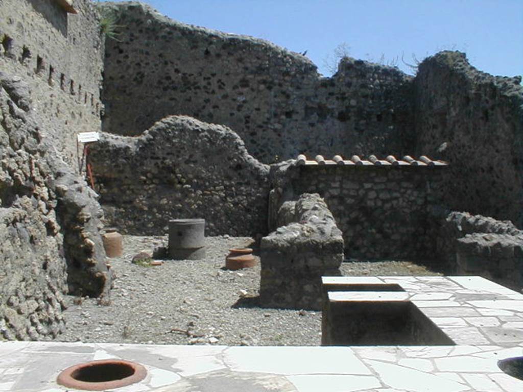 VI.16.40 Pompeii. May 2005. Room B, looking east across counter towards two rear rooms C and D and a corridor E to rear, leading to rooms F and G. According to NdS, room B was the bar-room, around which were smaller rooms. Adjacent to the north wall, near the entrance doorway, was the two-sided masonry sales counter or podium. At the other end in the centre of the bar-room, it ended with a kitchen oven (a), also in masonry. The external vertical surface of the sales counter was painted in red. The horizontal surface was decorated with embedded triangular slabs of marble and other shapes. Leaning against the north wall were the usual three small masonry steps, painted in red, to display the goods and housewares. The room was rather rustic.
At the east end of the north wall of the bar-room, near the doorway to room C, was a doorway to the area below the stairs at VI.16.39. Rooms C and D were rooms for the use of the clients of the bar. When excavated, the entrance doorposts on the left showed the imprint left by the timber. At the north end of room G, behind room C, was the latrine (b).
