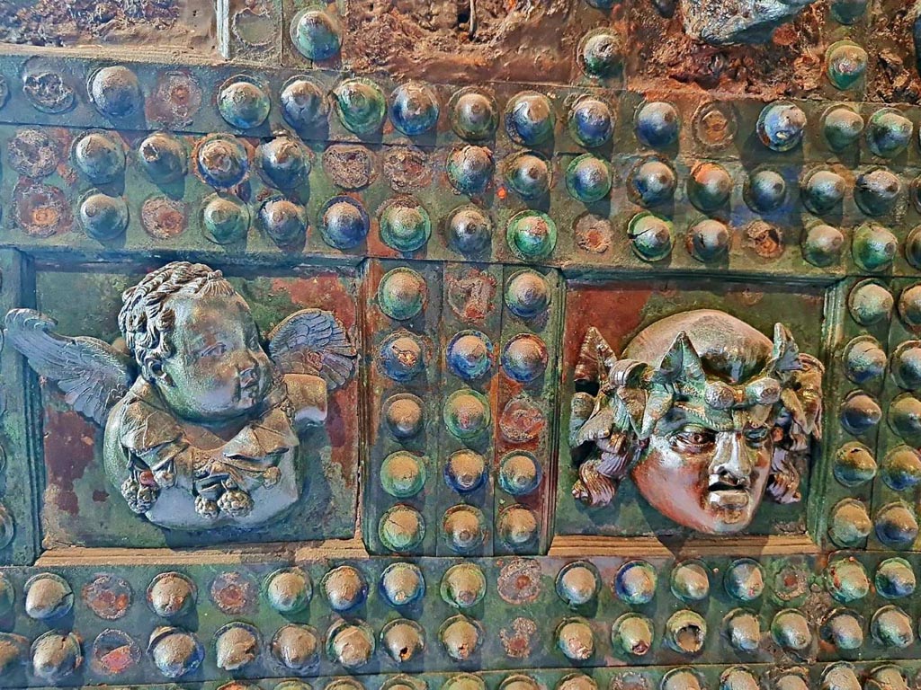 VII.2.18 Pompeii. October 2023.
Arca/strong box, detail front of box –
(on left) winged cupid carrying a garland of vine leaves and grapes around the neck, and (on right) a mask with long hair crowned with ivy.
Photo courtesy of Giuseppe Ciaramella.