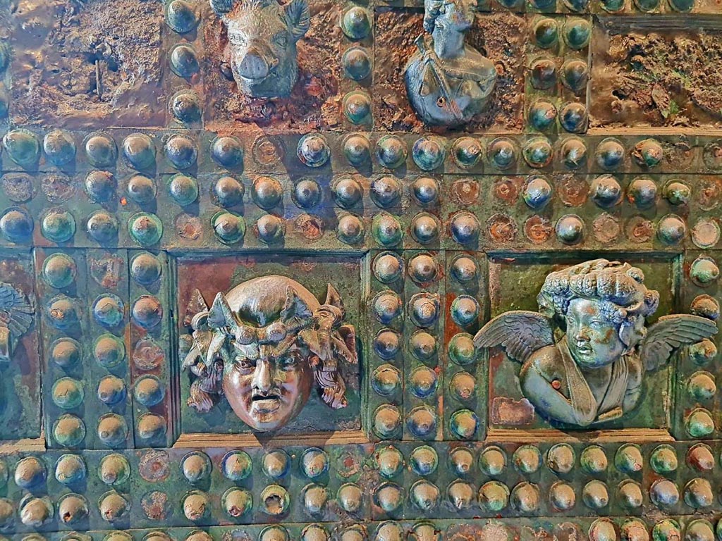 VII.2.18 Pompeii. October 2023.
Arca/strong box, detail from front of box –
(on left) a mask with long hair crowned with ivy, and (on right) the head of a winged cupid richly crowned with grape leaves.
Photo courtesy of Giuseppe Ciaramella.