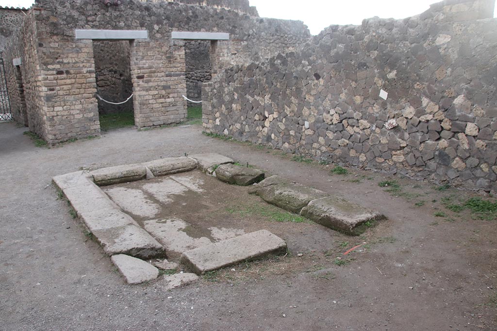 VII.7.2 Pompeii. October 2024.
Looking south across impluvium “g” made of slabs of tufa, from south side of tablinum “k”.
The doorway to room “d” is on the south side, with doorway to room “i” visible behind the wall. Photo courtesy of Klaus Heese.