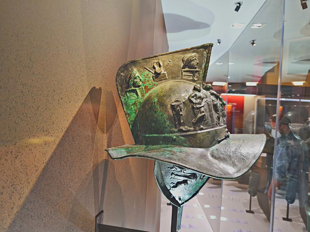 VII.11.6 Pompeii. Found 28th March 1863.
Photo taken May 2021 in display at Naples Archaeological Museum. Photo courtesy of Giuseppe Ciaramella.
According to the description card –
“The bronze and silver helmet with a depiction of Muses, the bronze greave with cupids and the fragments of a rectangular shield which were found together at Pompeii in March 1863, they may plausibly have been part of the armour of a murmilla.”