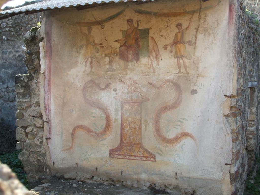 VII.12.11 Pompeii. December 2004. Lararium. According to Boyce, on the west wall was a lararium painting on a white background. Upon a throne sat Vesta, on each side of the goddess stood a wreathed Lar, much smaller than Vesta. Behind and to the right of her throne, the forepart of an ass was visible. In the lower zone were two serpents approaching a cylindrical altar painted in imitation yellow marble and finished with fire and fruit. Each serpent had a red crest and beard, but those on the right serpent were larger. At the sides of both the upper and lower panels were green trees and above the top of the upper panel were two garlands. The entire painting was bordered with broad red stripes at top and sides. Boyce thought this one of the most finely executed of all lararium pictures.
See Boyce G. K., 1937. Corpus of the Lararia of Pompeii. Rome: MAAR 14. (p.70, no.316, and Pl. 24,1)