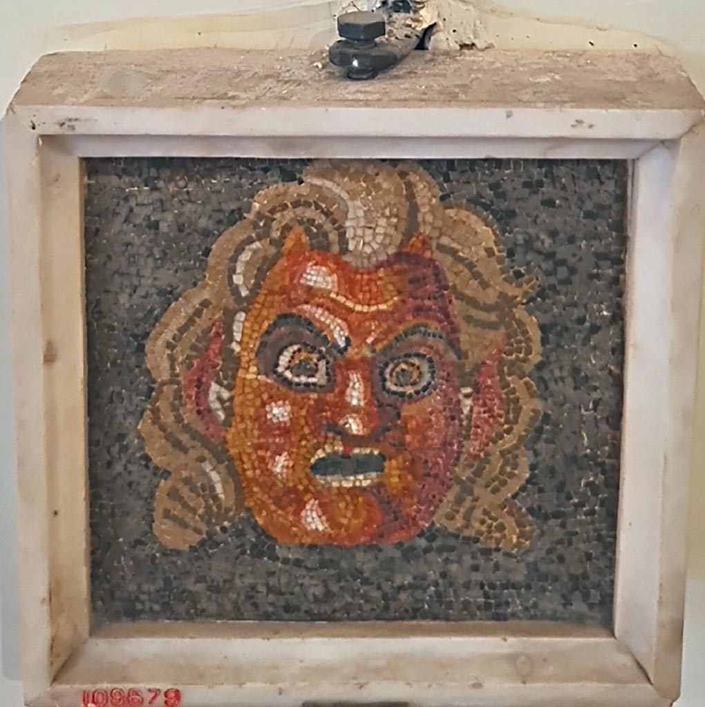 Mosaic mask found at VII.13.23 on 17th November 1821.
Now in Naples Archaeological Museum. Inventory number 109679.
According to the Giornale, in this location were found two beautiful masks in mosaic embedded above two stones of marble.
See Fiorelli G., 1864. Pompeianarum antiquitatum historia, Vol. 3: Naples, p. 30, dated 17th November 1821.
According to the information card –
Theatrical mask.
Fragment of a mosaic pavement in opus vermiculatum made from polychrome tesserae on a black background, depicting a theatrical mask of Pan. (Early Ist century BC.)
(Maschera teatrale.
Frammento di mosaico pavimentale in opus vermiculatum di tessere policrome su fondo nero, raffigurante una maschera teatrale di Pan.
Inizi del I secolo a.C.)
Photo courtesy of Giuseppe Ciaramella. September 2024.