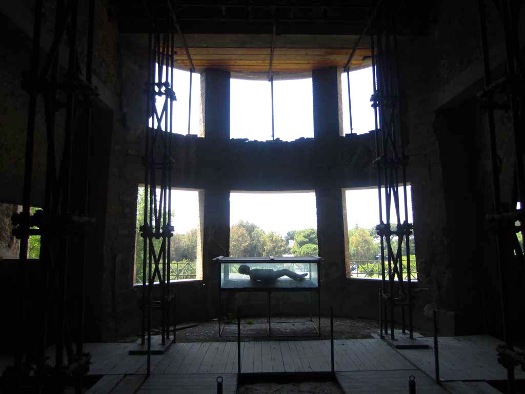 VII.16.17-22 Pompeii. May 2012. Oecus 62, looking west towards curved apse with six windows, in oecus.
A plaster cast made of a body is on display in the case. Photo courtesy of Marina Fuxa.
