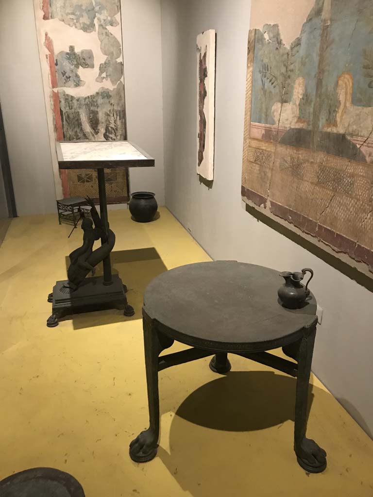 VII.16.17-22 Pompeii. April 2019. On display in Antiquarium, from House of Fabius Rufus.
Bronze table with cupid riding a dolphin and marble top. PAP inventory number 13371.
Bronze table with lion's feet. PAP inventory number 13108.
On table is a bronze pitcher with face of a follower of Dionysus. PAP inventory number 14072.
Photo courtesy of Rick Bauer.