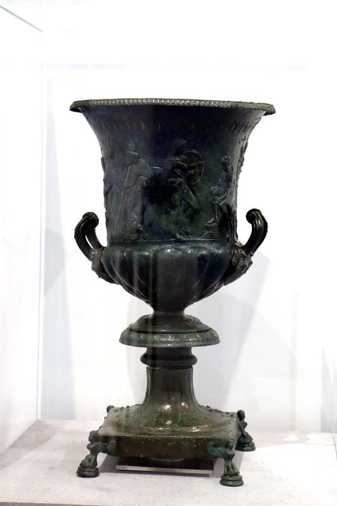VIII.1.4 Pompeii. February 2021.
Bronze krater with scenes in relief, found in the triclinium at IX.13.1-3, on display in Antiquarium.
Photo courtesy of Fabien Bièvre-Perrin (CC BY-NC-SA).
According to the description card in the Antiquarium -
The bronze krater was decorated with eight armed male figures, six standing and two sitting.
One of the seated figures is tying his shoelaces.
A young man stands in front of him holding a shield, with a bearded figure holding a staff behind.
Young and naked, the second seated figure caresses an animal with one hand and holds a sword by the hilt in the other.
(See IX.13.1-3 for more photos).