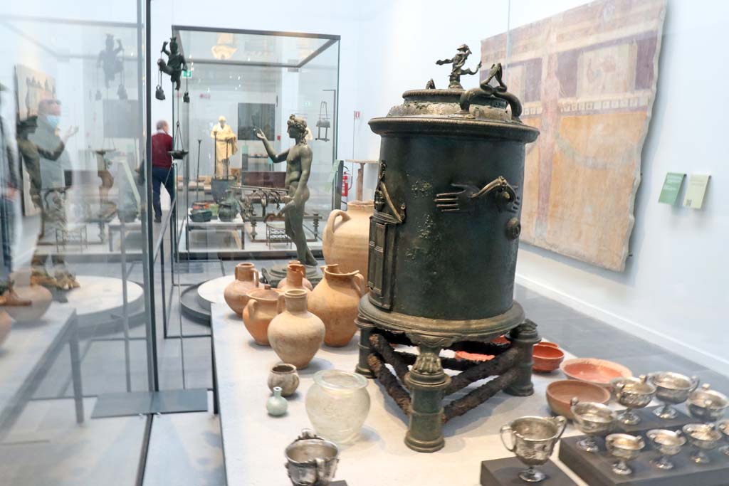 VIII.1.4 Pompeii. February 2021. 
Looking across display cabinet containing silver from Moregine and bronze food warmer from I.8.17 (this end), and at the far end – 
Bronze strainer and two pitchers from new excavations in Reg. V,
and Terracotta bottles, jugs and red ware plates and bowls from room 11, of the House of Orion, V.2.
Looking towards the bronze Ephebus lamp holder from VII.16.19-22, and across to the marble statue of Nike, from Oplontis, at the far end.
On the wall to the right is part of a painted wall from I.6.2.
Photo courtesy of Fabien Bièvre-Perrin (CC BY-NC-SA).

