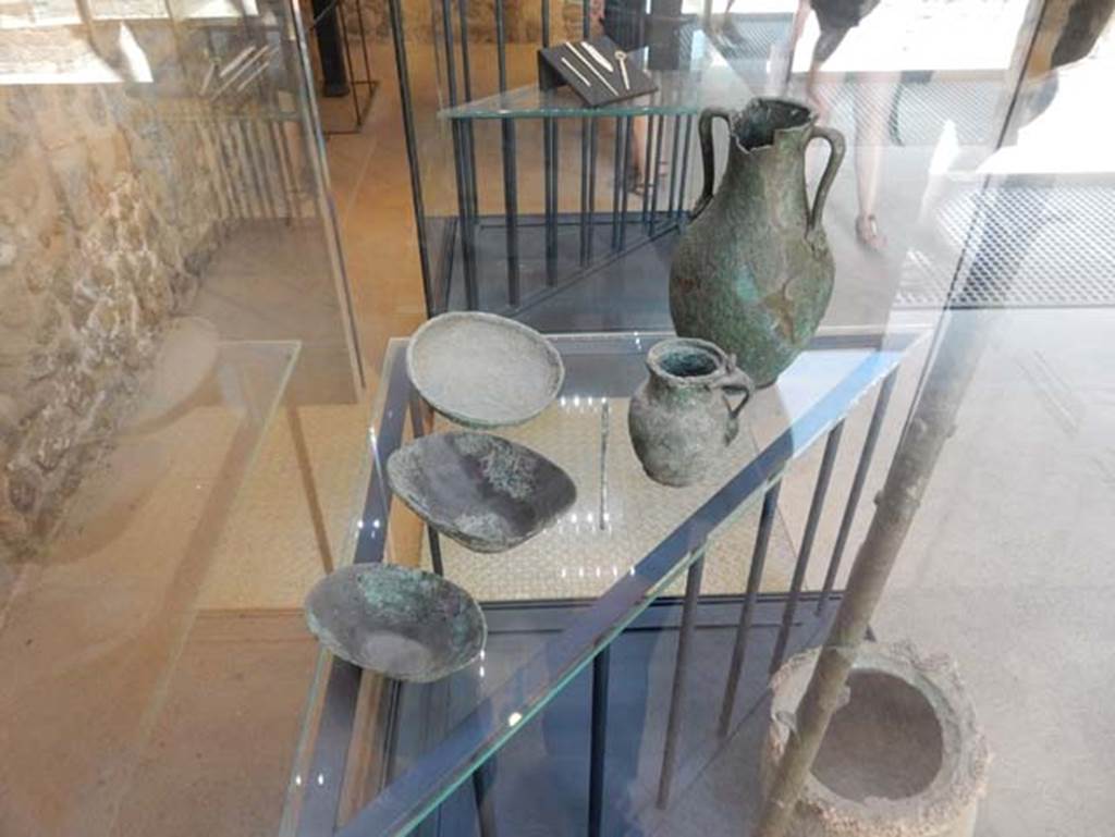 VIII.2.1 Pompeii. May 2018. Finds from excavations conducted in the area since the 1930’s.
The top shelf includes
A bronze needle. Pompeii inventory number 6312.
Bronze jug with female face at lower joint of the handle. Pompeii inventory number 6319.
Small bronze amphora decorated with theatre masks at the lower joints of the handles. Pompeii inventory number 6321.
Three bronze cups, bronze needle, bronze jug with female face and small bronze amphora decorated with theatre masks. Pompeii inventory numbers 6310A, 6310B, 6323.
All were found in the area to the south of the House of Championnet.
Photo courtesy of Buzz Ferebee.