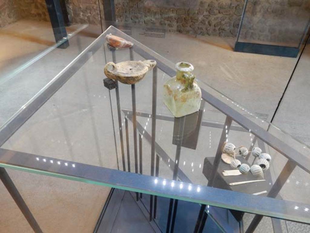 On display in Championnet complex Pompeii. May 2018. Finds from excavations conducted in the area since the 1930’s.
Elements of a glass paste necklace. Pompeii inventory number 6330.
Green glass bottle. Pompeii inventory number 14828.
Two terracotta lamps.
Found in the area south of the House of Championnet.
Photo courtesy of Buzz Ferebee.