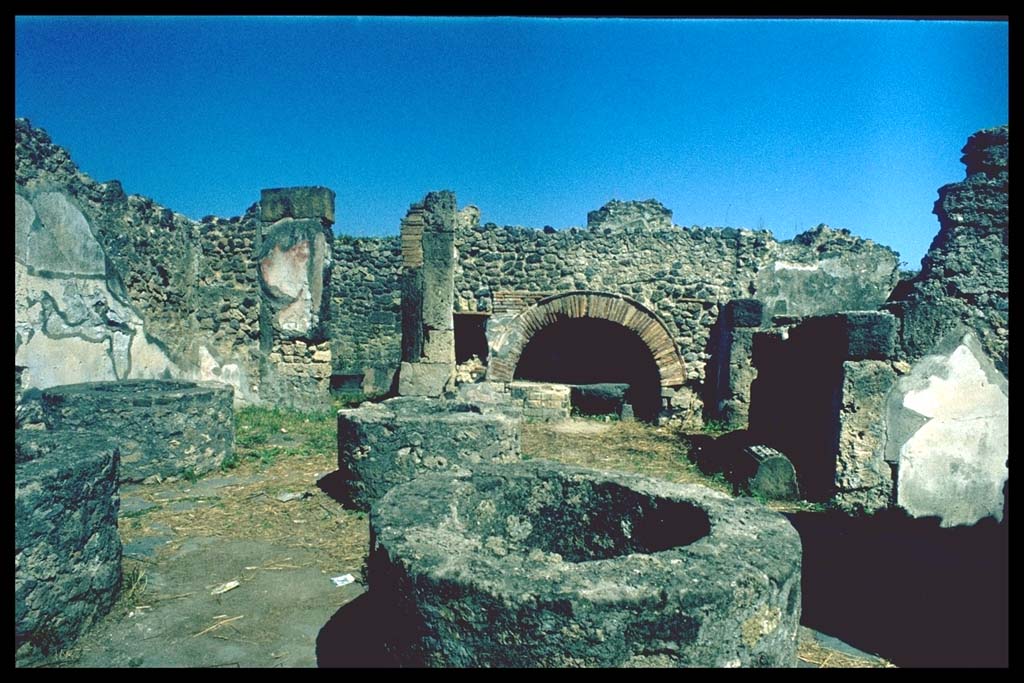 VIII.6.11 Pompeii. Bakery, looking east.
Photographed 1970-79 by Günther Einhorn, picture courtesy of his son Ralf Einhorn.
According to Mau, from the entrance corridor one immediately entered the bakery, where one could see the substructures of four mills, and around them the flooring for the mules.
The walls were painted simply in the last style, with red and yellow compartments.
Near to the north wall was a square stone that was probably used as a table.
A larger table was against the left wall (west), between the two doorways and was supported by two feet of wood, while the other side was supported by two beams fixed into the wall. The table cannot be currently recognised other than by these two beam holes (height 0.75, distance between them 2.10m) and the two holes in the floor (0.80 distance from the wall) where the wooden feet stood.
A similar table but smaller (1.m) was against the same wall between the doorway to “f” and the front corner (south-west corner).
“a” is the oven, (round, not elliptical, as it seems on the plan); before the oven on the left buried in the ground was a vessel for water, and next on the left was a large dolium.
See Bullettino dell’Instituto di Corrispondenza Archeologica (DAIR), 1884, p.138
(Per la fauce, che ha la porta immediatamente alla strada,s'entra nel pistrinum, ove troviamo le sostruzioni per quattro mulini, e intorno ad esse il selciato per i muli. Le pareti son dipinte semplicemente nell'ultimo stile, con compartimenti rossi e gialli. Addosso al muro N sta una pietra quadra di tufo che serviva probabilmente da tavola. Una tavola più grande stava addosso al muro sinistra (0), fra le due porte, ed era sorretta da due piedi di legno, mentre l'altro lato era con due travi fissato nel muro. Così presentemente la tavola non si riconosce che dai buchi di queste due travi (a. 0,75, distanti fra loro 2,10) e da due buchi corrispondenti nel pavimento, (distanti dal muro 0,80), ove stavano i piedi di legno. Una tavola simile ma più piccola (m. 1) stava addosso alla stessa parete fra la porta di “f" e l'angolo anteriore. – “a” è il forno (tondo, non ellittico, come sembra nella pianta); avanti ad esso sta a sinistra un vaso per acqua murato nel suolo, e accanto a sinistra un gran dolium.)
