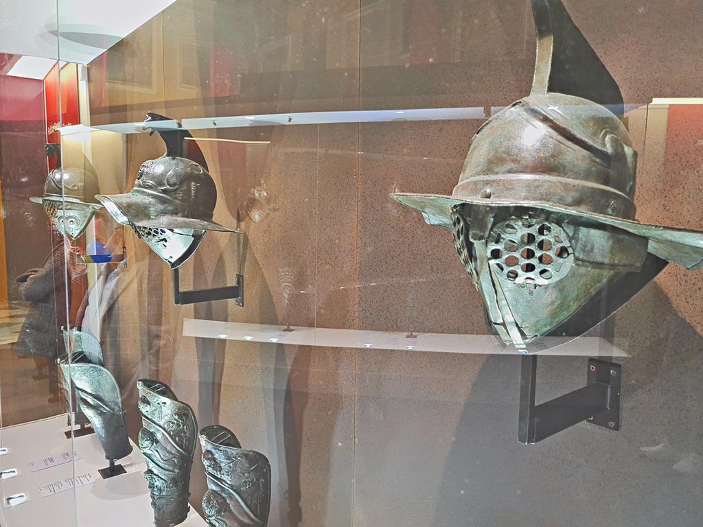 Naples Archaeological Museum display case of helmets and greaves. Photo taken May 2021, courtesy of Giuseppe Ciaramella.