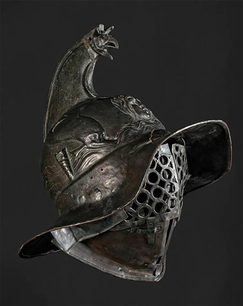 VIII.7.16 Pompeii. Thraex gladiators helmet.
The crest of this helmet ends in a griffin’s head and feathers could also be attached to it.
On the front, the helmet is decorated with a gorgon’s head, possibly Medusa.
On the sides, there are dolphins, as well as tubes into which feathers could be inserted.
The gladiator’s face was covered by a two-part visor.