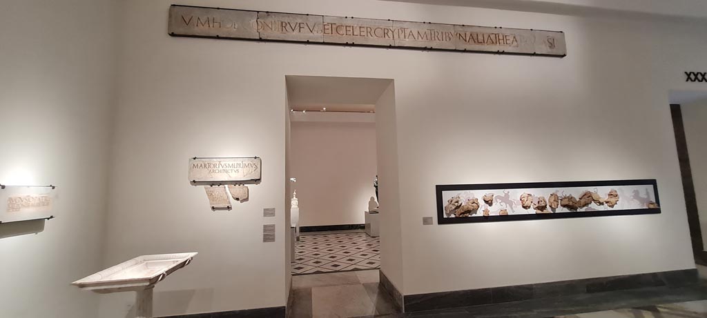 VIII.7.20 Pompeii. April 2023.
Above doorway, (inv. 3821) one of the two identical white marble slabs with inscriptions each well over 6 metres long, from the area of the stage.
Left side of doorway, two marble slabs (inv. 3834 and 3823).
No. 3834 provides the name of the architect involved in the refurbishment of the theatre – M. Artorius Primus freedman of Marcus.
No. 3823 is a white marble slab in two parts, stating that M. Holconius Rufus and M. Holconius Celer arranged at their own expense for the enlargement of the seating and tribunal of the theatre and the construction of a vaulted corridor.
On display in “Campania Romana” gallery in Naples Archaeological Museum. Photo courtesy of Giuseppe Ciaramella.