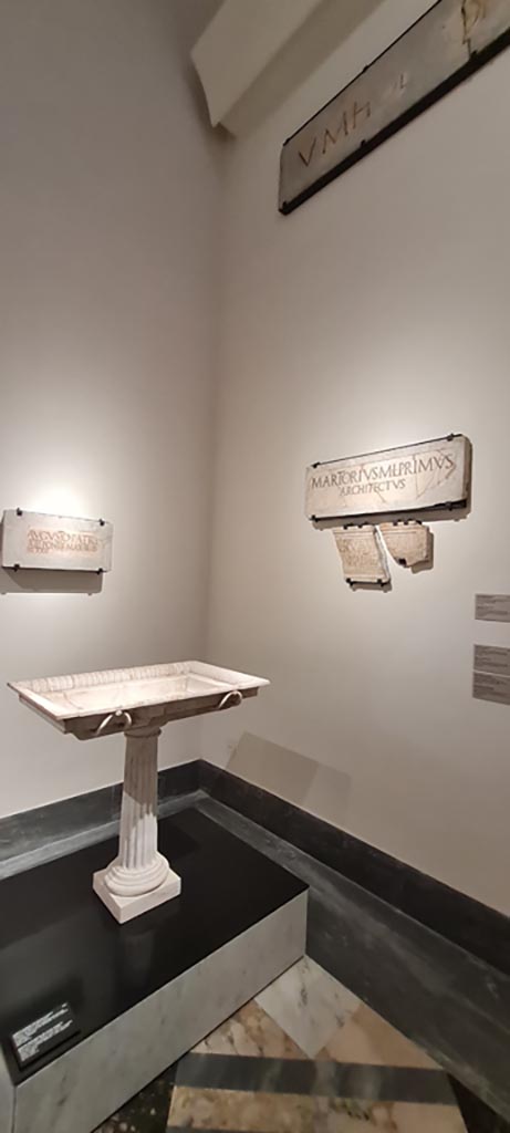 VIII.7.20 Pompeii. April 2023.
White marble basin of a fountain (inv. 6868) on display in “Campania Romana” gallery in Naples Archaeological Museum.
On left, Dedication to Augustus,
On right, marble slabs ((inv. 3834 and 3823).