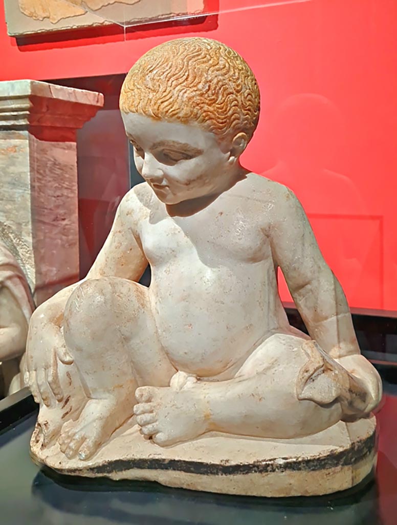 IX.12.9 Pompeii. March 2024. Fountain statuette of a boy and dolphin. Marble 1st century AD.
(Statuetta di fontana con bambino e delfino. Marmo, l sec. D.C.)
Provenanced from this house according to the information card at the exhibition entitled -
“L’altra Pompei, vite comuni all’ombra del Vesuvio”. Photo courtesy of Giuseppe Ciaramella.
According to an information card in the house –
“The garden was also decorated with a fountain which was crowned with a statue of a young boy, on which we can identify traces of original colours.”