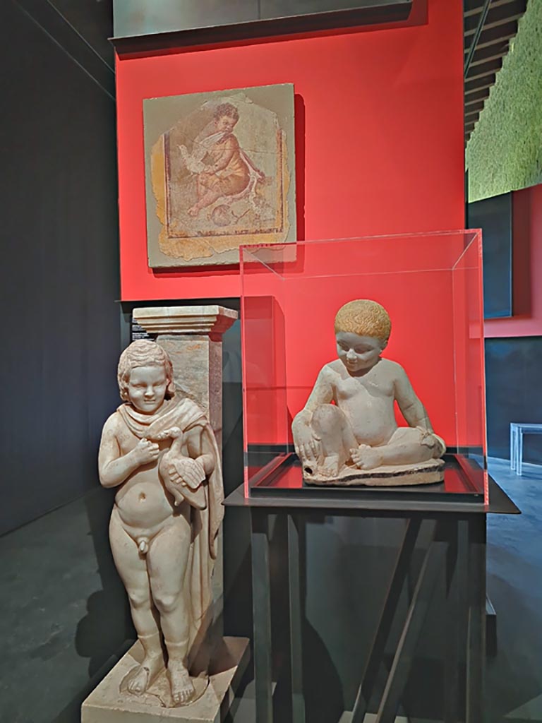 March 2024.
Photographed at the exhibition in the Palaestra entitled “L’altra Pompei, vite comuni all’ombra del Vesuvio”.
Fresco, at top – Adonis as a child, from I.9.3, House of Successus,
On the left – Monopodium with boy holding a goose, from I.9.3, House of Successus,
On the right – Fountain statuette of a boy and a dolphin, from IX.12.9, House of the Painters at work.
Photo courtesy of Giuseppe Ciaramella.